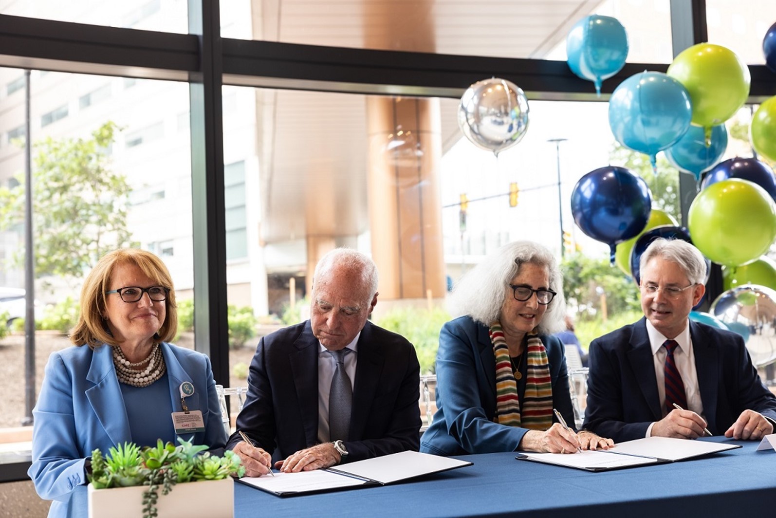 Group of four adults seated at a table signing documents with blue, green, and silver colored balloons in the background.